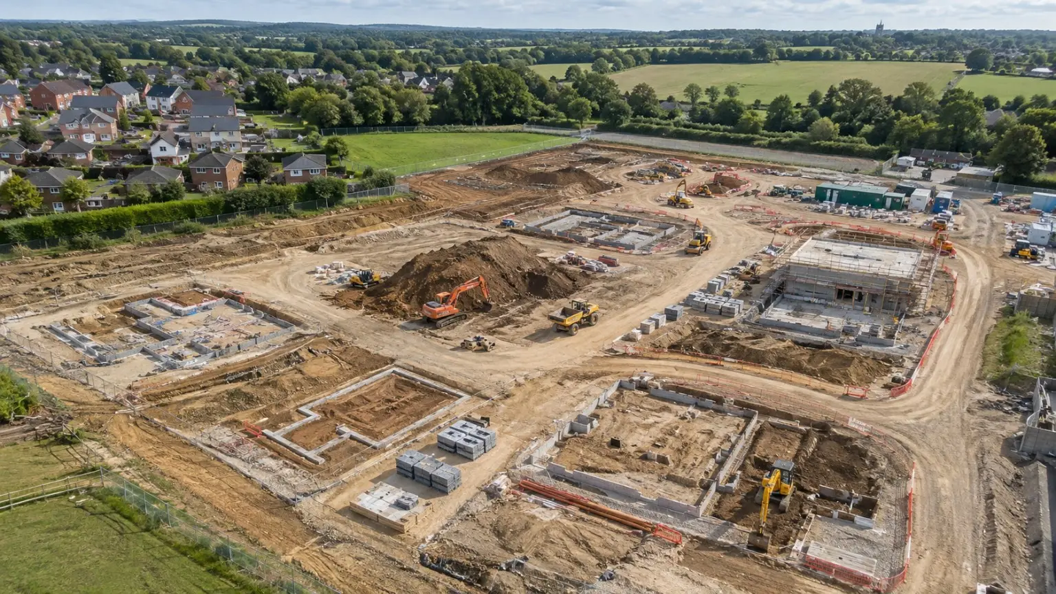 Aerial view of a housing development at groundworks stage with earthworks, foundations, machinery and surrounding countryside